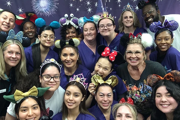 Group of smiling people wearing colorful mouse ear headbands, posing together in front of a festive backdrop.