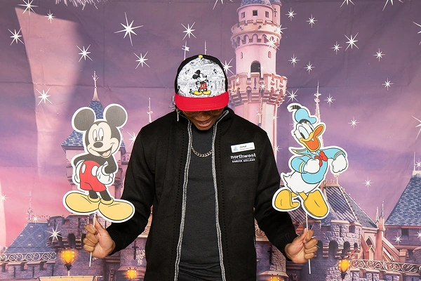 Person holding cutouts of Mickey Mouse and Donald Duck in front of a castle backdrop, wearing a cap with Mickey design.
