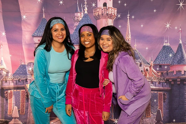 Three women in colorful matching outfits and headbands pose together in front of a castle backdrop.