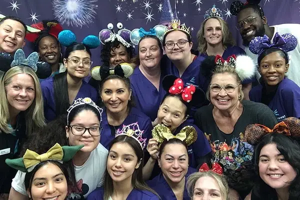 Group photo of diverse individuals wearing colorful mouse ear headbands, smiling in front of a festive backdrop.