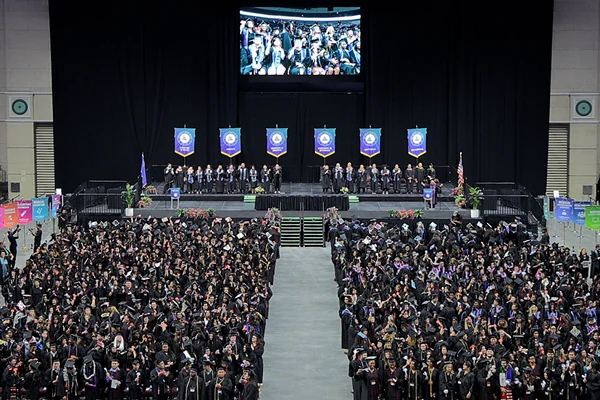 A large crowd of graduates in caps and gowns celebrates at a graduation ceremony, with a stage and banners in view.