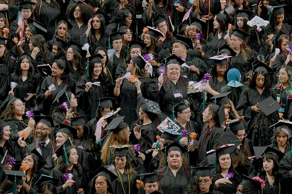 A large group of graduates in black caps and gowns, celebrating with flowers and smiles at a graduation ceremony.
