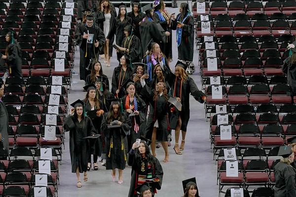 Graduates in black gowns walk through a stadium filled with empty chairs, celebrating their achievement.