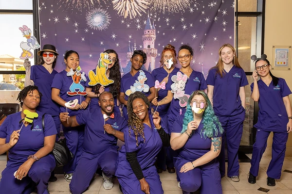A group of 11 people in purple scrubs pose with cartoon character props against a festive backdrop.