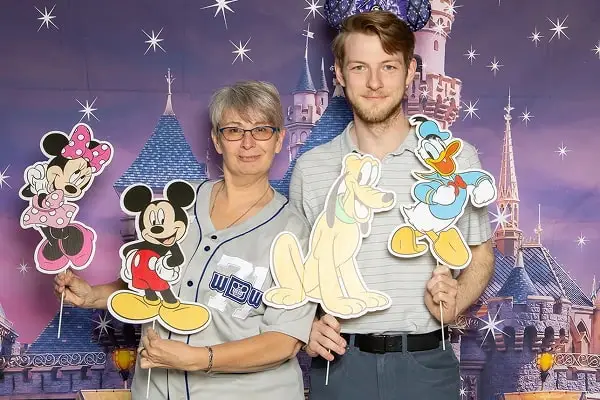 A woman and a man hold cartoon character cutouts in front of a castle backdrop, smiling at the camera.