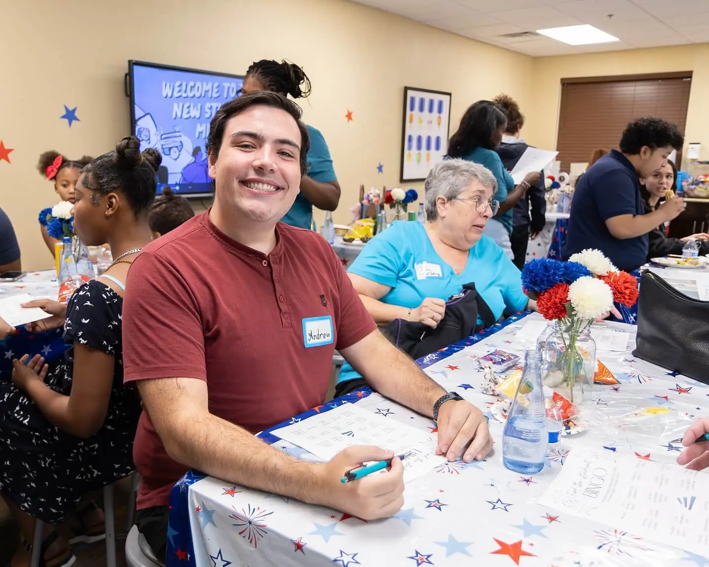 A smiling man sits at a table with others, surrounded by decorations and snacks, during a community event.