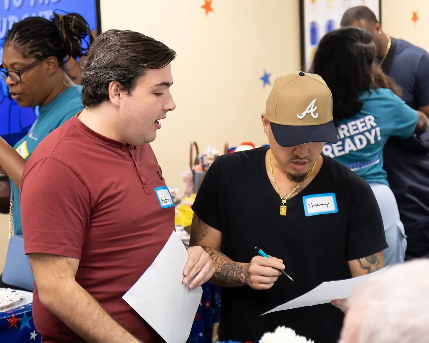 Two young men discuss a paper in a lively room with colorful decorations and people in the background.