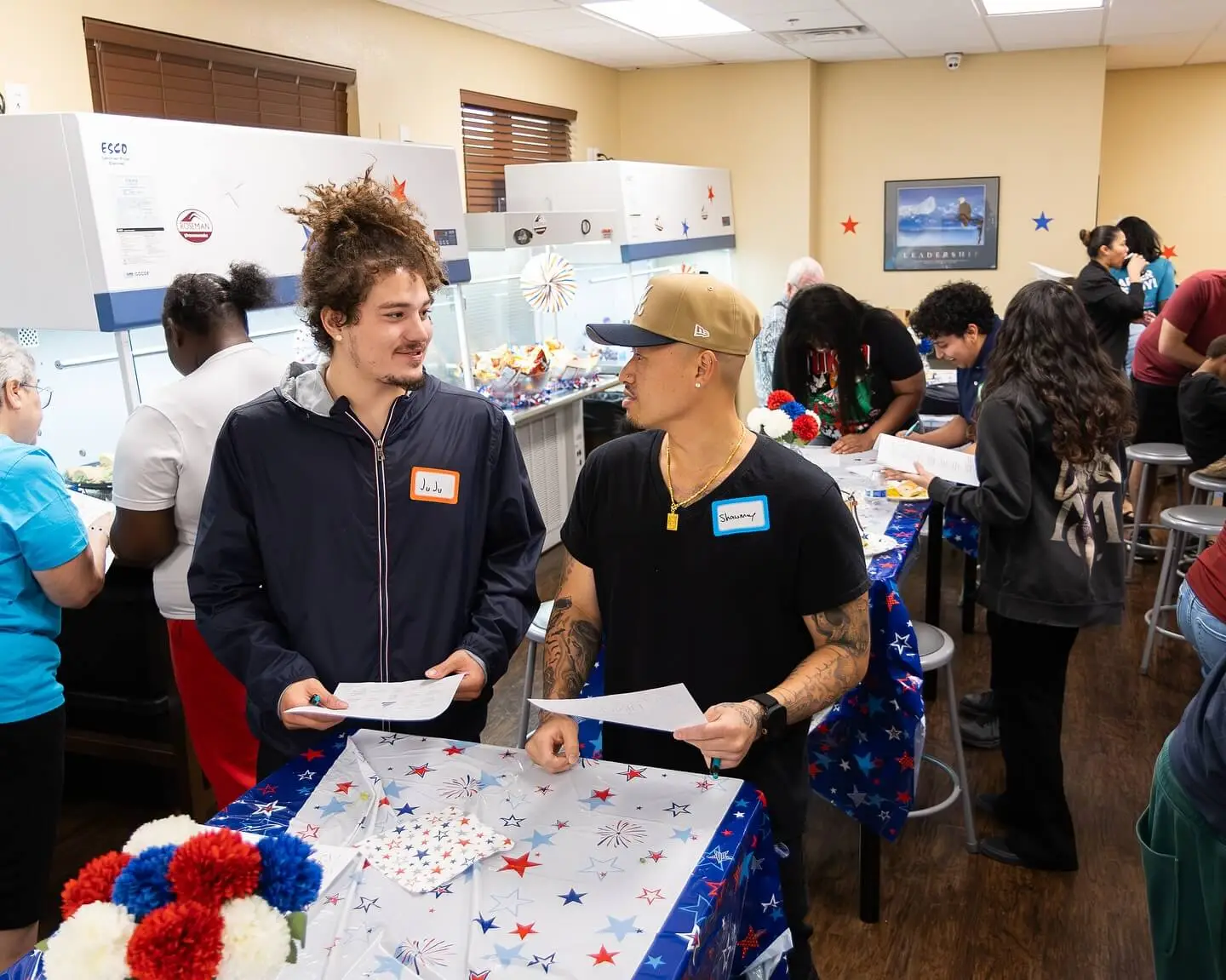 Two young men talk at a decorated table in a busy community space, with others engaged in activities around them.
