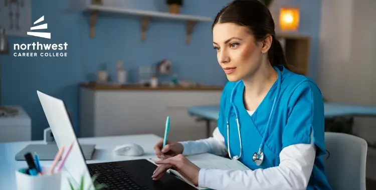 A focused student in scrubs studies at a laptop, taking notes in a bright, modern classroom setting.