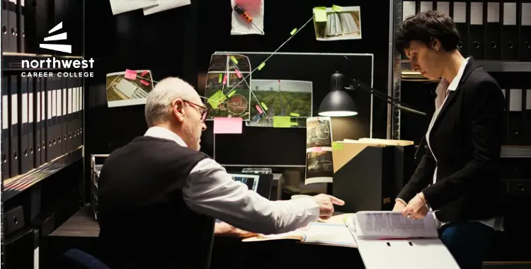A man and woman discuss documents at a desk, surrounded by files and notes in a dimly lit office.