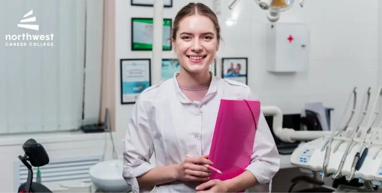 Smiling female dental assistant in a clinic, holding a pink folder, with dental equipment and certificates in the background.