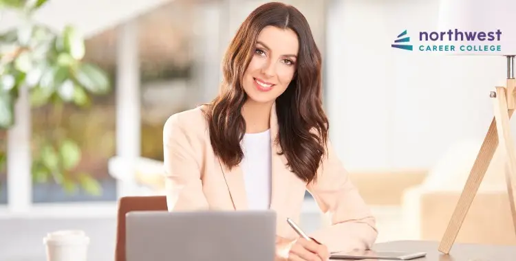 A smiling woman in a light blazer sits at a desk with a laptop, holding a pen, in a bright, modern office setting.