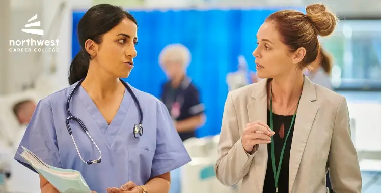 Two women, one in scrubs and the other in a blazer, engage in conversation in a healthcare setting.