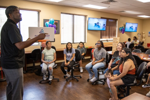 A speaker addresses a group of engaged youth and adults in a classroom setting, with balloons and screens in the background.