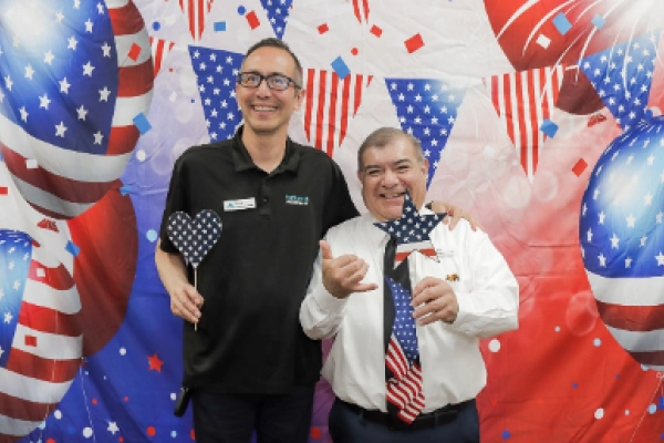 Two men pose together with festive props against a colorful backdrop of red, white, and blue decorations.