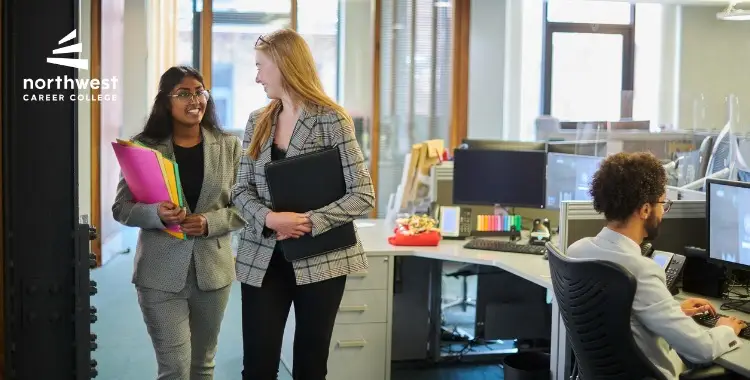 Two women in professional attire walk through an office, chatting and holding folders, while a man works at a desk.