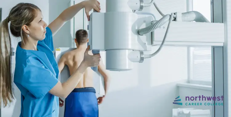 A healthcare professional adjusts an X-ray machine while a patient stands with their back turned, preparing for imaging.