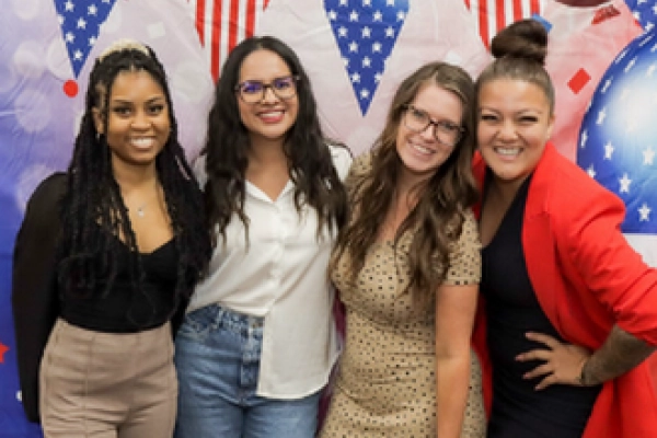 Four women smiling together in front of a festive backdrop with red, white, and blue decorations.