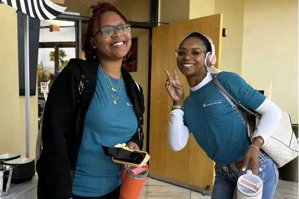 Two smiling women in matching blue shirts pose together, one making a peace sign, in a bright indoor setting.
