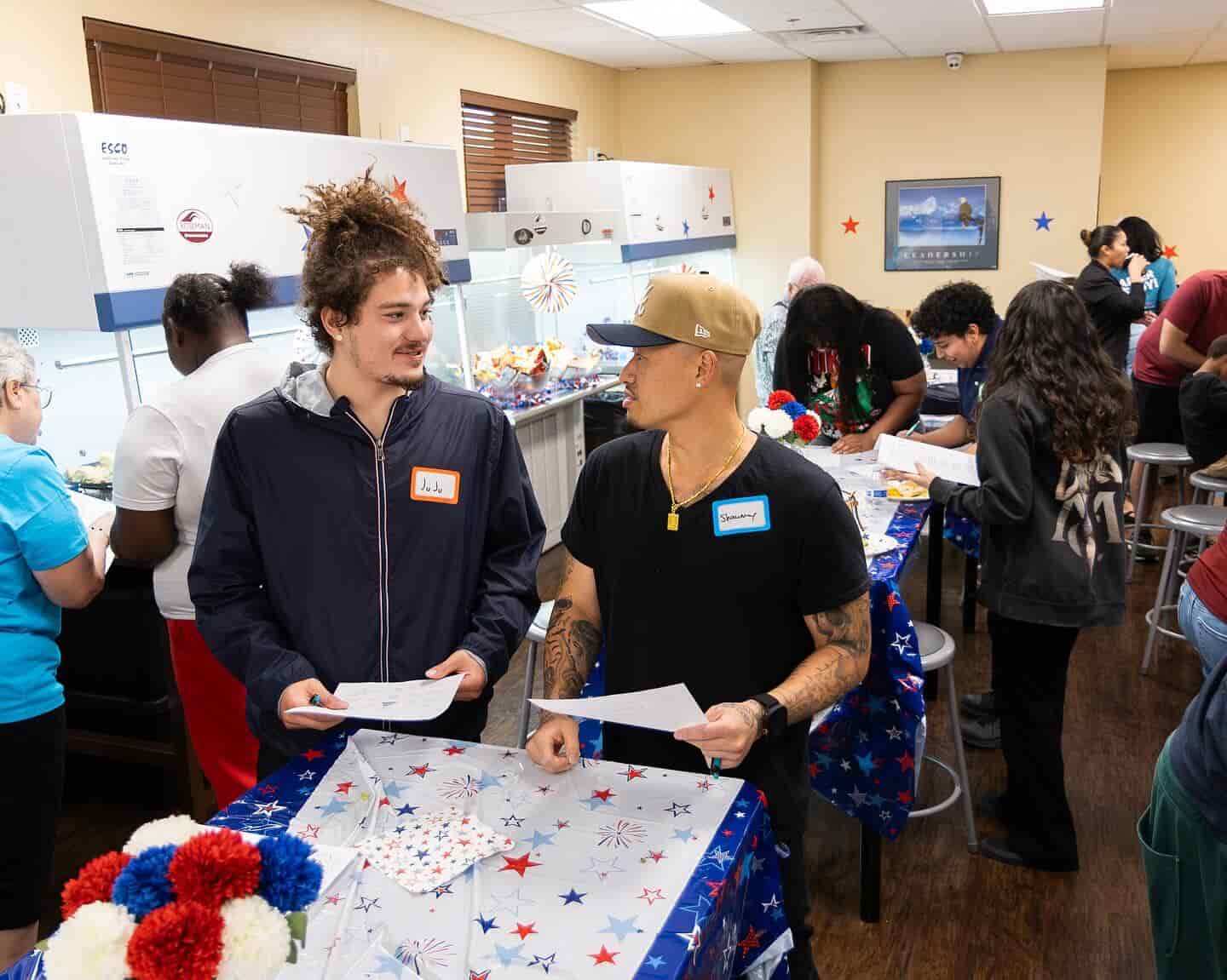 Two men chat at a decorated table while others work in a lively community kitchen setting.