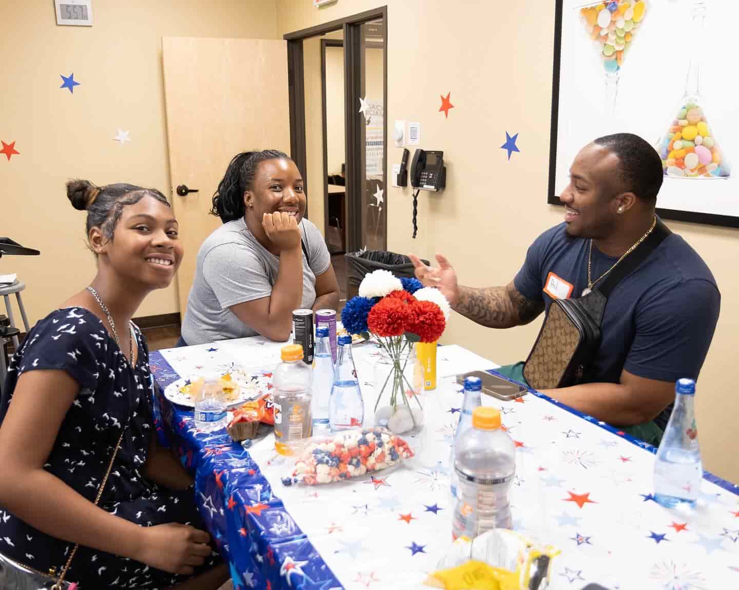 Three people sit at a decorated table, smiling and chatting, with snacks and drinks around them.
