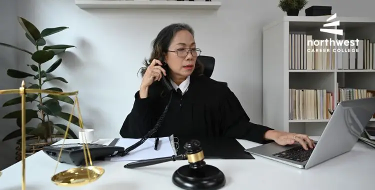A judge in a black robe speaks on the phone while working at a desk with a laptop and gavel.