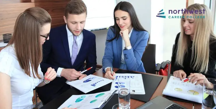 Four professionals collaborate at a conference table, discussing charts and graphs with pens and papers in hand.