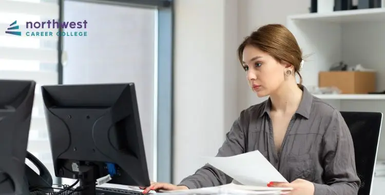 A woman sits at a desk, focused on a computer screen while holding papers, in a bright office setting.