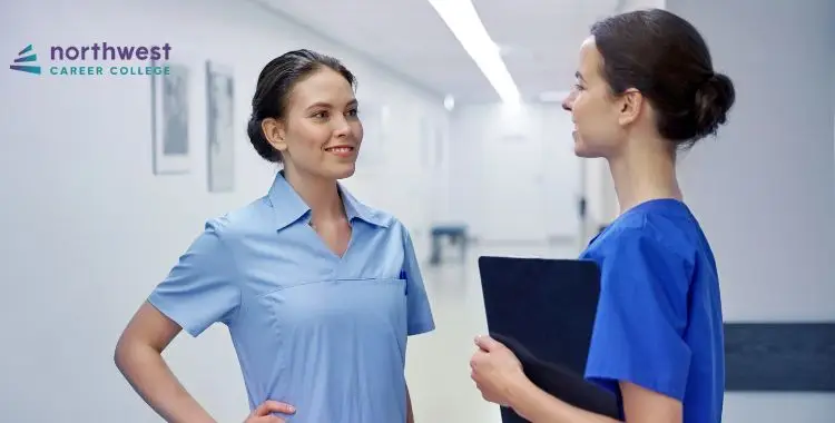 Two healthcare professionals in scrubs converse in a well-lit hallway, with a logo for Northwest Career College in the.