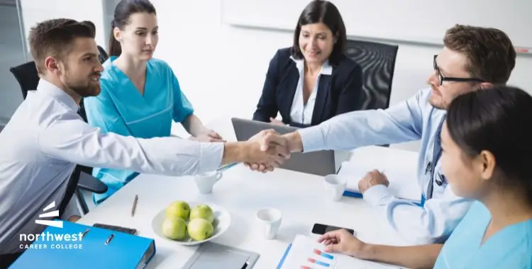 A group of professionals in a meeting, shaking hands and discussing, with documents and apples on the table.