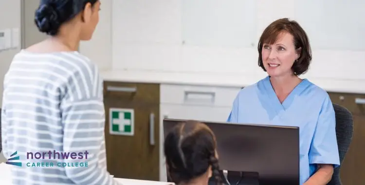 A healthcare professional in scrubs speaks with a woman at a reception desk, with a child visible in the background.
