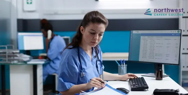 A healthcare professional in scrubs reviews documents at a computer station in a modern office setting.