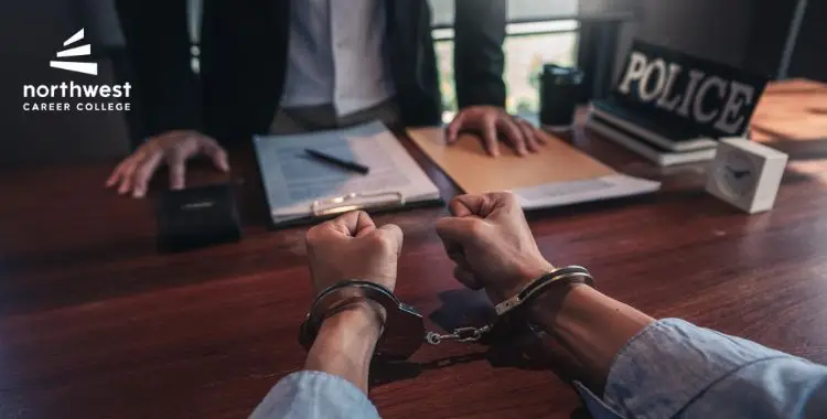 Hands in handcuffs on a table with police documents and a person in a suit in the background.