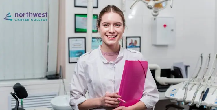 Smiling woman in a dental office holding a pink folder, wearing a lab coat, with dental equipment in the background.