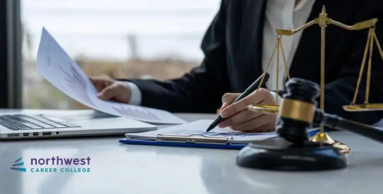 A person in a suit reviews documents and takes notes beside a laptop and a gavel on a desk.