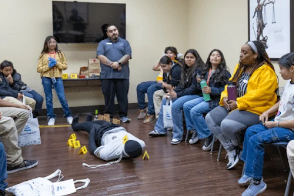 A group of children observes a mock crime scene with a dummy and evidence markers in a classroom setting.