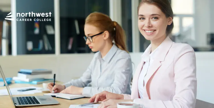 Two women working at a desk; one smiles at the camera while the other writes notes. A laptop and books are visible.