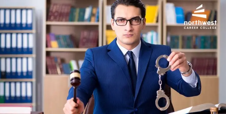 A serious man in a suit holds a gavel and handcuffs, seated in a library setting with legal books.