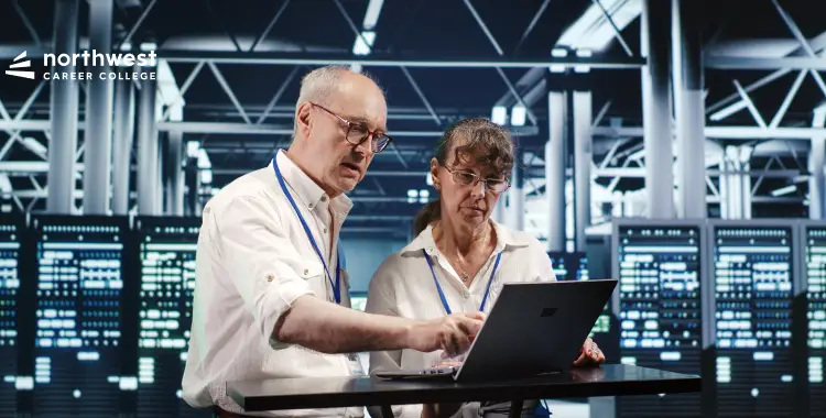 Two professionals collaborate at a laptop in a tech environment, with server racks in the background.