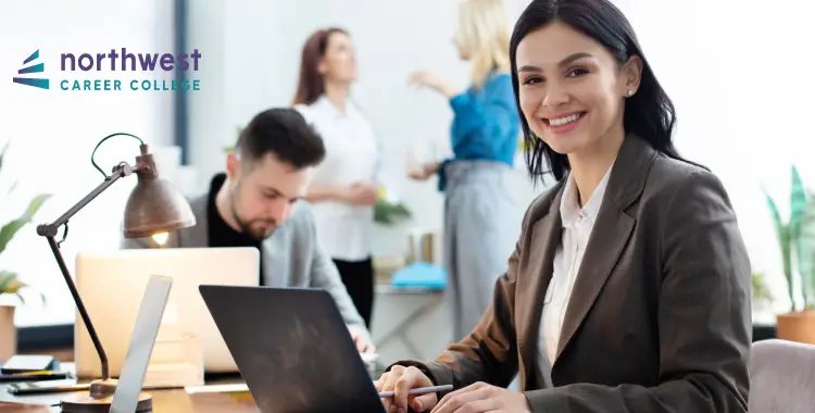 A smiling woman in a suit works on a laptop in a bright office with colleagues in the background.