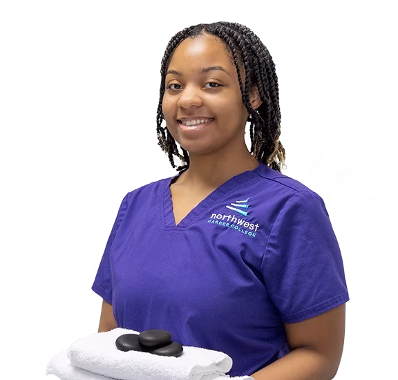 Smiling student in a purple uniform holds hot stones and a towel, representing massage school Las Vegas.