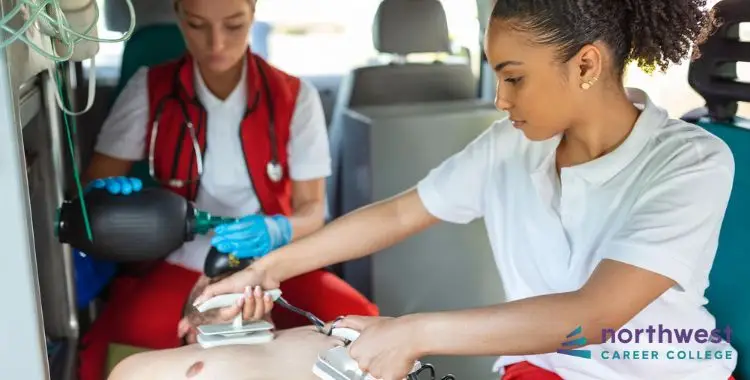 Two paramedics in an ambulance; one uses a defibrillator while the other prepares equipment.