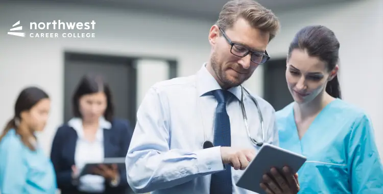 Two healthcare professionals discuss information on a tablet in a clinical setting, with others in the background.