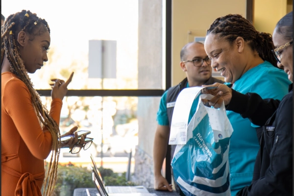 A woman in orange talks to healthcare workers at a reception desk, holding a bag and a document.