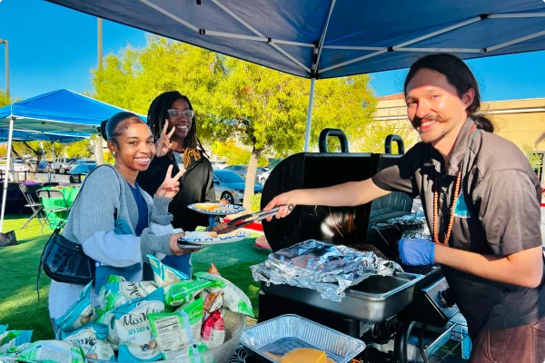 A man with a mustache smiles as he serves food from a grill using tongs to two female students at the "New Student Orientation: Summer-4C260803 August" event. The students smile and hold plates, one giving a peace sign. In the foreground, there is a large bowl filled with bags of potato chips, and the scene is set outdoors under a blue canopy tent.