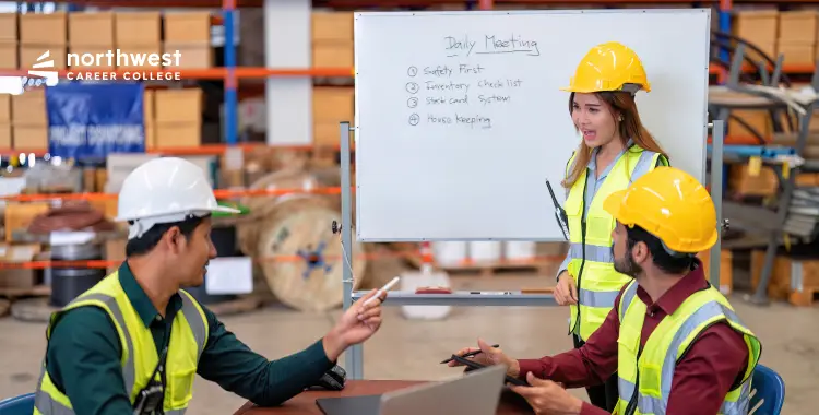 A woman leads a daily meeting with two men in safety vests and helmets, discussing a whiteboard list in a warehouse.