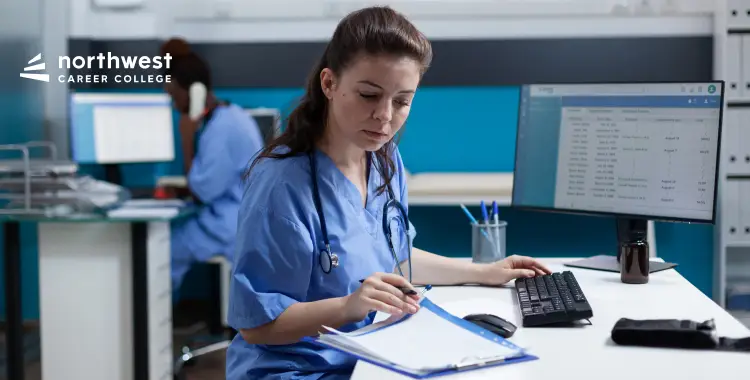 A healthcare professional in scrubs reviews notes at a desk with a computer, focused on her work.