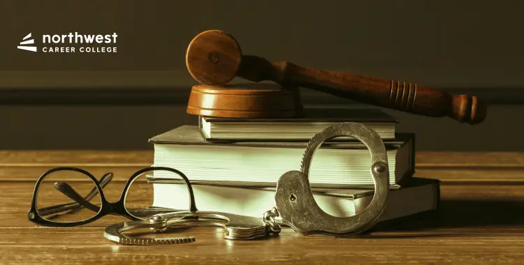 A gavel, handcuffs, glasses, and books arranged on a wooden surface, with a college logo in the corner.