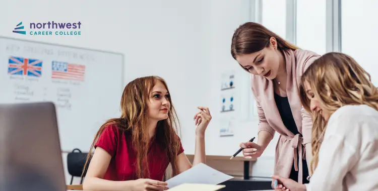 Three women collaborate at a table, discussing documents in a classroom setting with educational materials in the background.