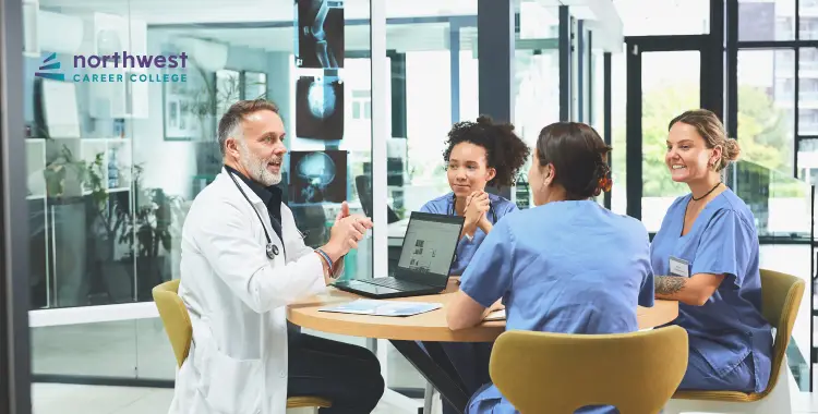 A doctor discusses with three nursing students around a table in a modern classroom setting.